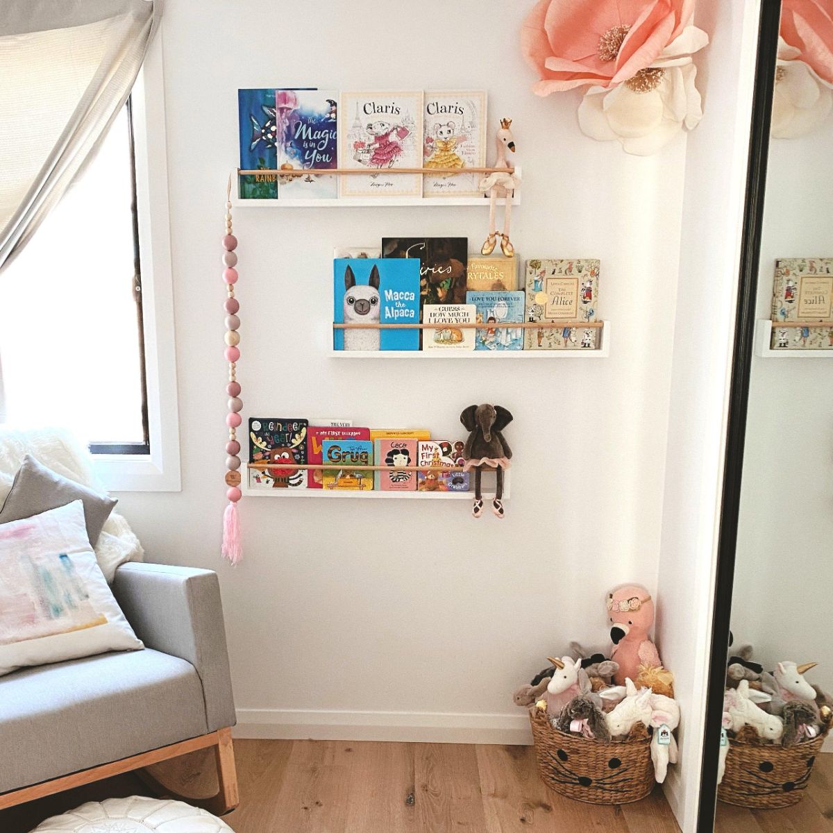 The wall has 3 white bookshelf with wooden peg across. The bookshelf has books and plush toys