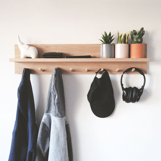 A long wooden floating entryway shelf with seven peg hooks mounted on a white wall. Two hoodies, a black cap, and wireless headphones hang from the pegs. The top shelf holds an elephant figurine, a wallet, a phone, and three small potted succulent plants.