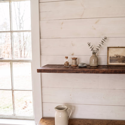 A rustic dark wood floating shelf mounted on a white shiplap wall, styled with small glass jars, a ceramic vase with eucalyptus, a stone, and a framed print. A white ceramic pitcher sits below on a lower shelf, next to a window looking out onto a wooded yard.