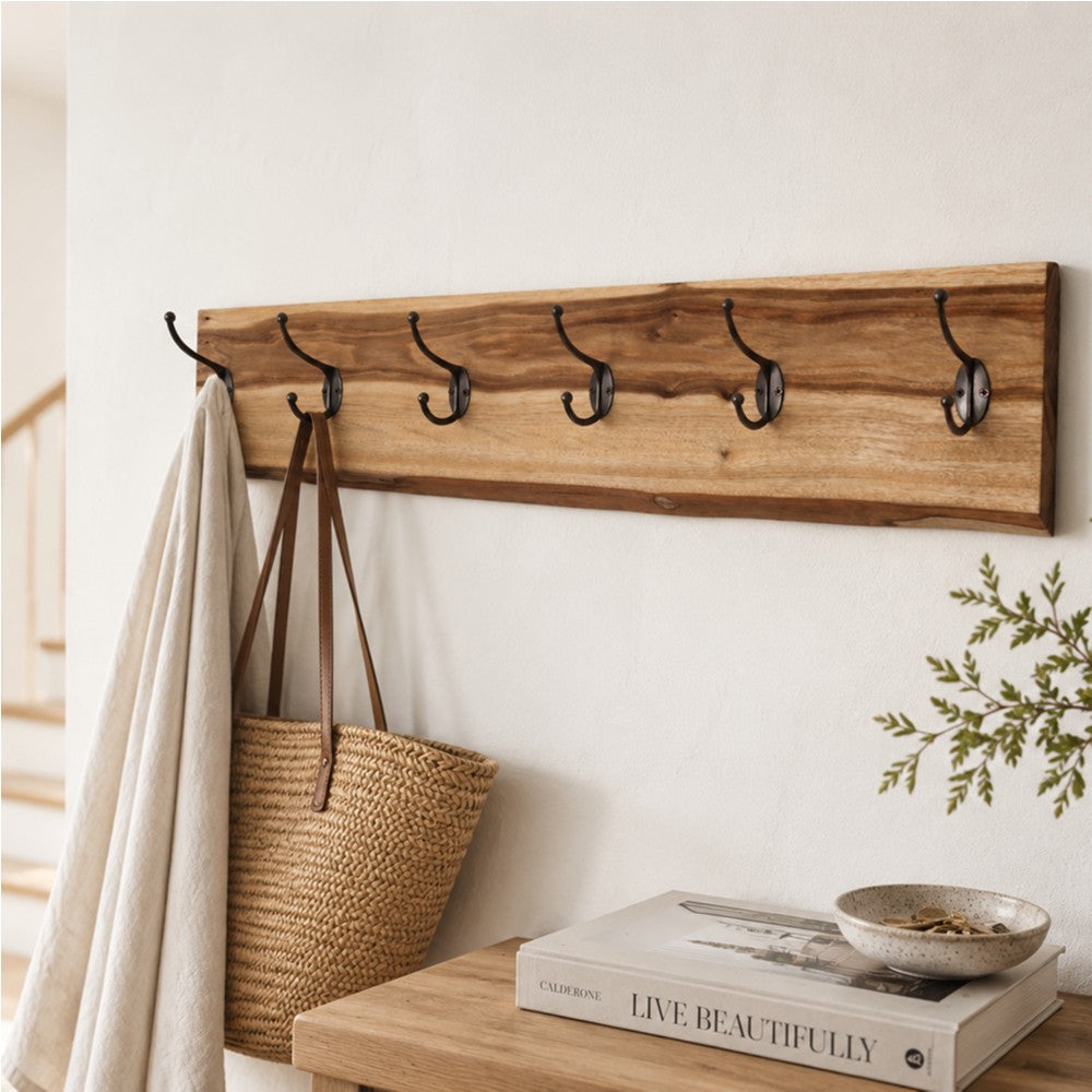 A straight-on view of a wall-mounted live-edge camphor laurel wood coat rack featuring six dark metal double hooks. A cream-colored linen cloth and a woven straw tote bag with leather straps hang from the hooks against a textured white wall.