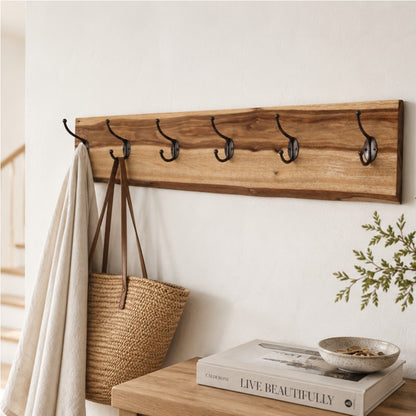 A straight-on view of a wall-mounted live-edge camphor laurel wood coat rack featuring six dark metal double hooks. A cream-colored linen cloth and a woven straw tote bag with leather straps hang from the hooks against a textured white wall.