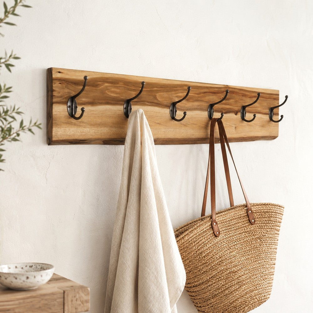 A left-angle perspective of a natural live-edge wooden coat rack with six black metal hooks mounted on a white wall. A linen towel and straw bag hang from the side, while a wooden console table 