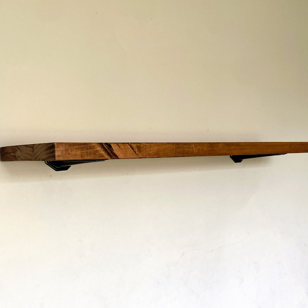 Side view of a long rustic pine floating shelf in walnut brown mounted on a plain white wall with black under-mount brackets.