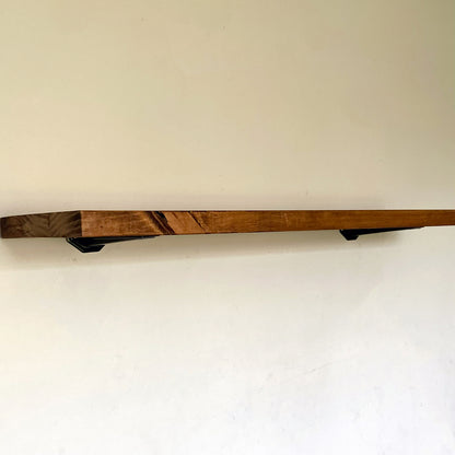 Side view of a long rustic pine floating shelf in walnut brown mounted on a plain white wall with black under-mount brackets.