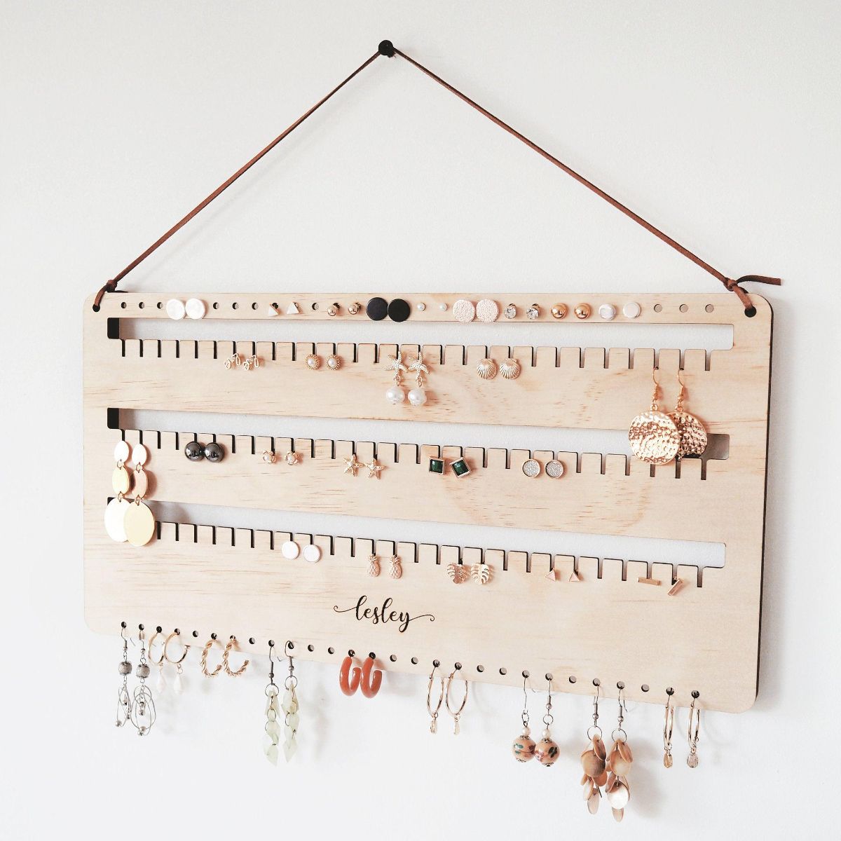 Left Side view of a personalised wall earring holder with leather string hanging on a screw. Stud, hook and dangling earrings are organised on this earring holder. A perfect gift for mum
