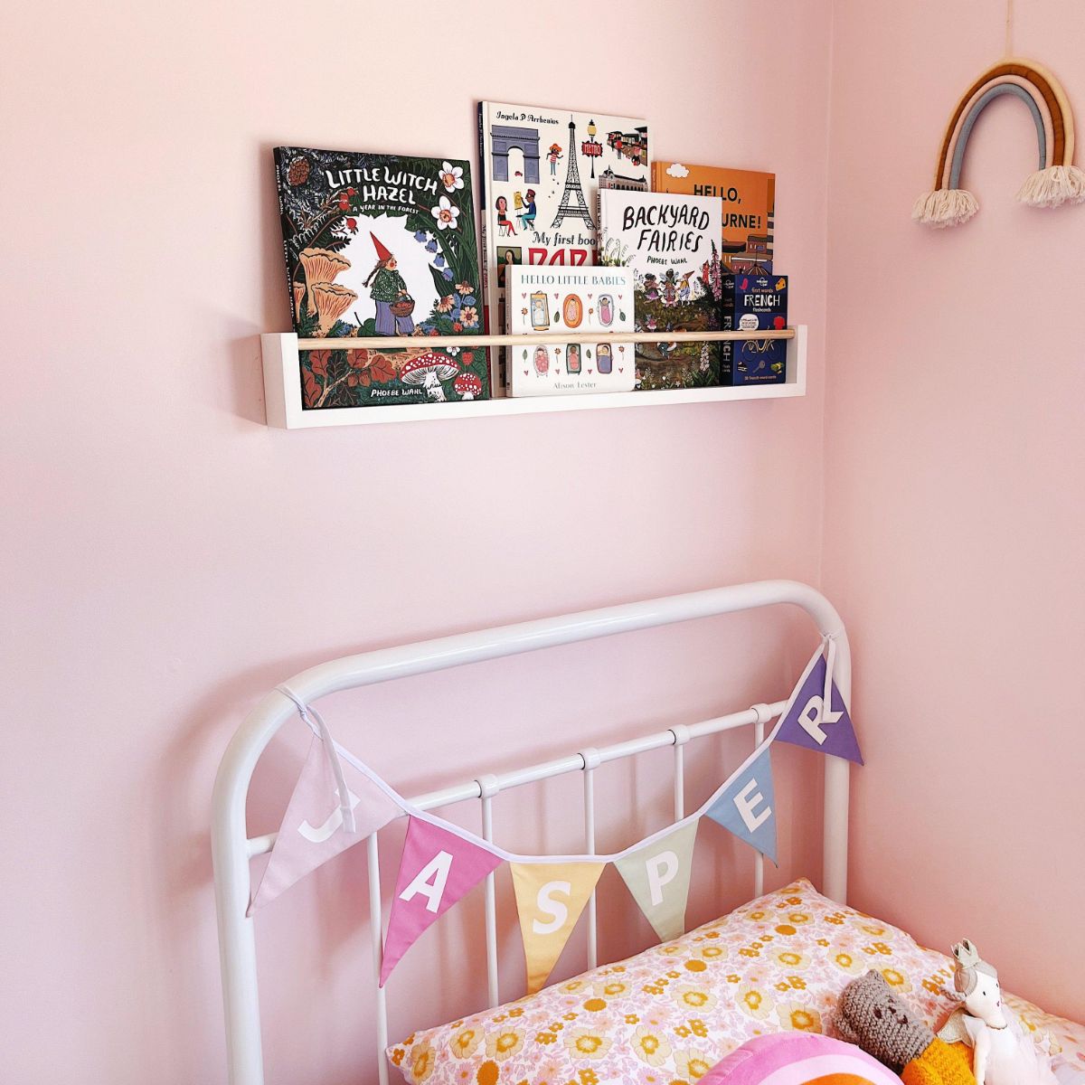 This is a 80cm white bookshelf with wooden peg across. It is placed on top of a bed with a pink wall as a background
