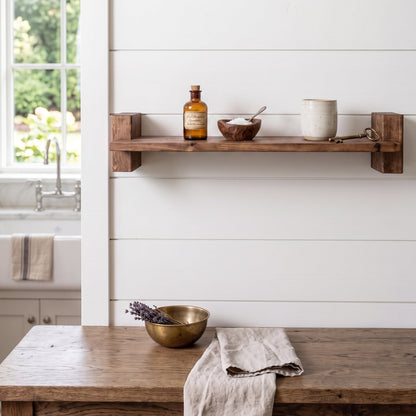 A rustic dark wood floating shelf mounted on a white shiplap wall, styled with an amber glass bottle, a small wooden bowl, and a ceramic mug. Below the shelf, a matching wooden table holds a brass bowl with lavender and a beige linen towel.