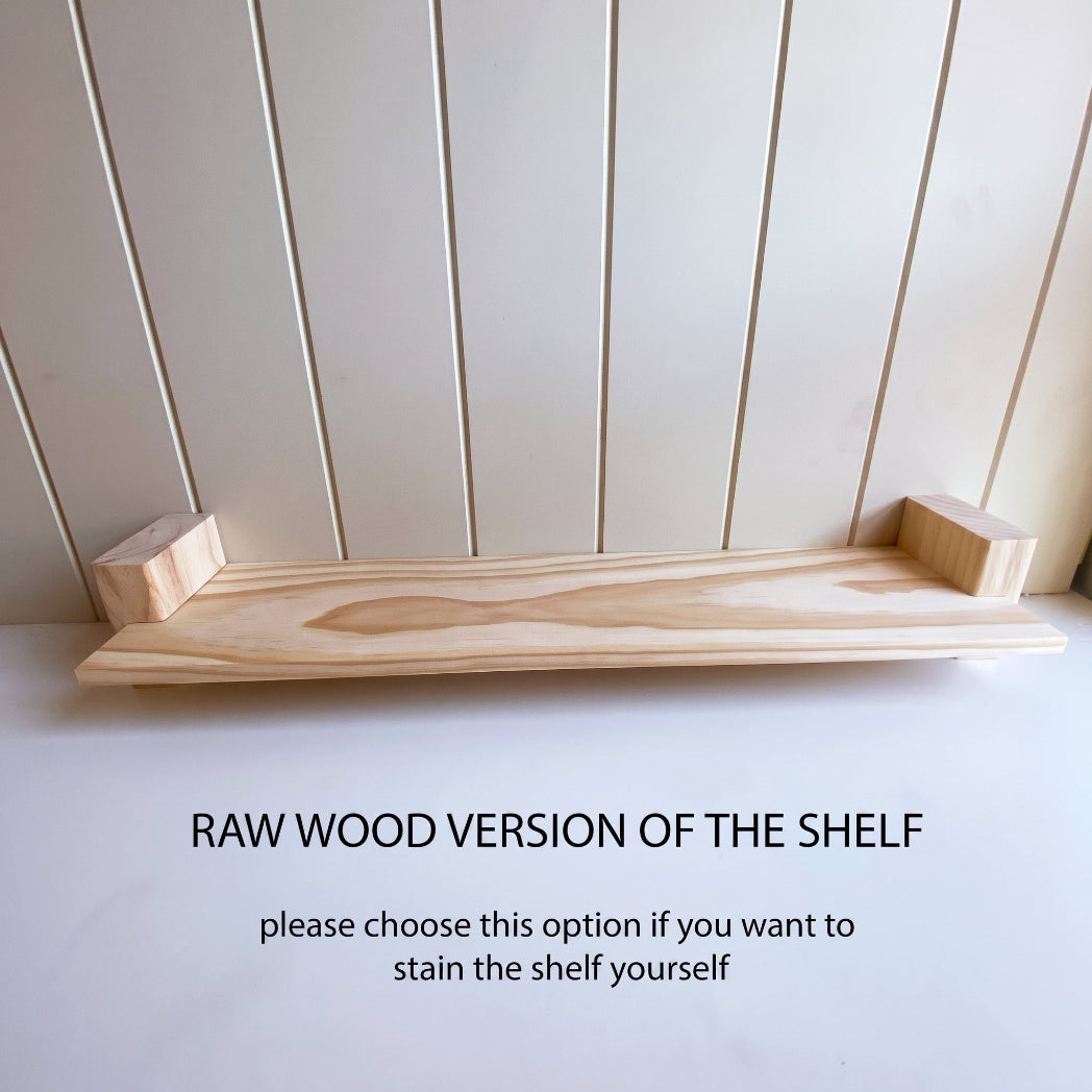 A raw wood floating shelf with two small end brackets, shown against a light beige paneled wall and white surface. Text below reads 'RAW WOOD VERSION OF THE SHELF' and 'please choose this option if you want to stain the shelf yourself'.