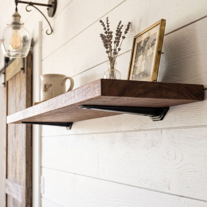 Close-up angled view of a handcrafted floating wooden shelf showing heavy-duty matte black metal brackets, mounted on a white shiplap wall in a farmhouse 