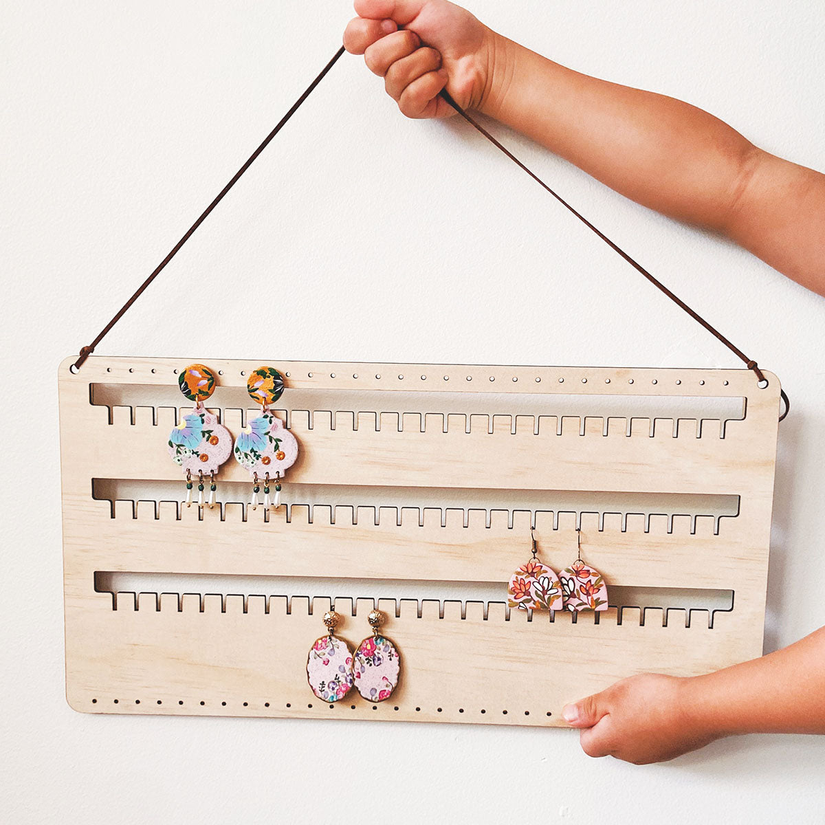 A small girl (full body not seen, only arms and hands) is holding a personalised wall earring holder with leather string hanging on a screw. Stud, hook and dangling earrings are organised on this earring holder.